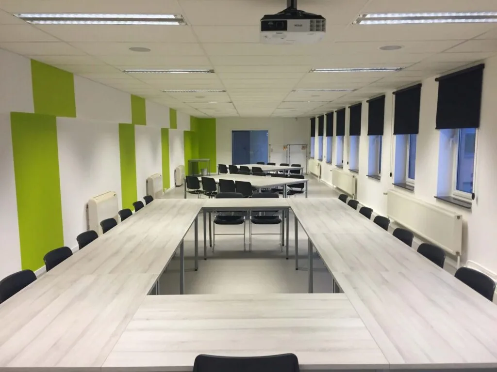 Modern conference room with U-shaped white tables, black chairs, and green and white walls.