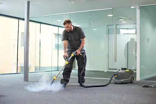 A professional cleaner performing carpet steam cleaning in an office space, using a steam cleaning machine that emits visible steam, ensuring a deep and hygienic clean.