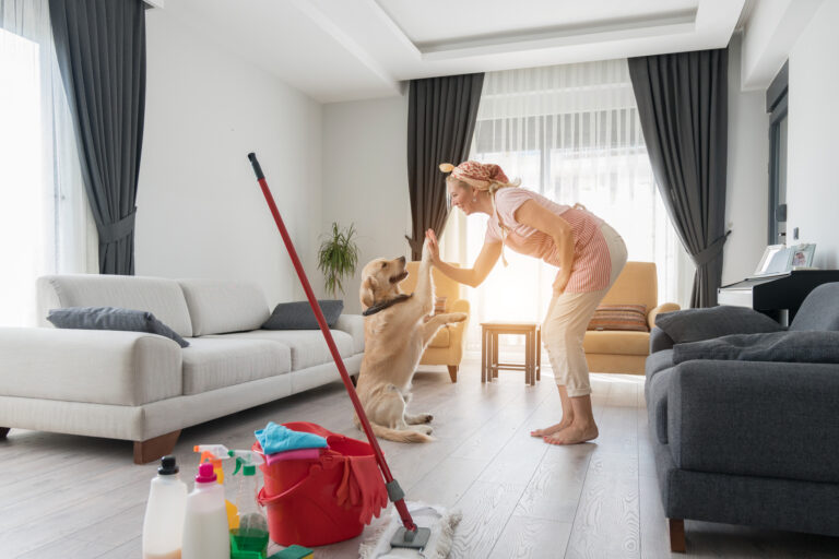 A happy cleaner high-fiving a client's dog in a spotless living room after house cleaning, with cleaning supplies visible in the foreground.