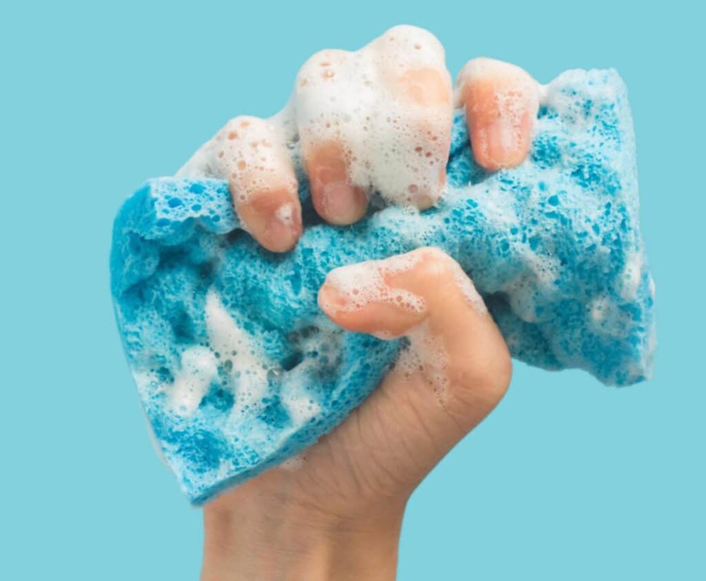 A hand squeezing a blue soapy sponge against a plain turquoise background